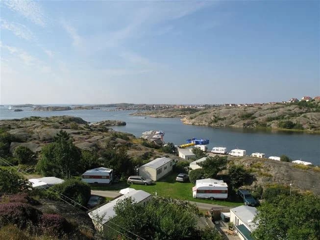 En översiktsbild av Wiggersviks Camping med en parkering i förgrunden, båtar förtöjda vid en marina längs kusten, och natursköna vyer över ett sjödistrikt i bakgrunden.