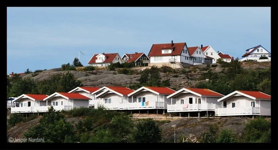 Bild av Wiggersviks camping med en rad sommarstugor i en lugn, grönskande miljö. Stugorna har röda fasader, vita knutar och traditionella sadeltak. I förgrunden syns verandor med utemöbler.