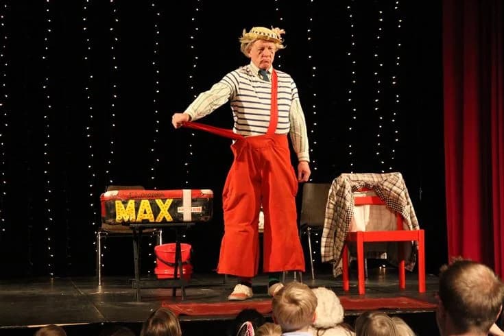 En teaterscen med ett pågående framträdande på Wiggersviks camping. Artister i kostymer står på scenen framför en röd ridå och uppträder för publiken.