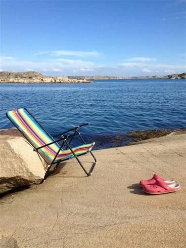 Utsikt över en solig strand vid Wiggersviks camping med klarblå himmel och glittrande hav. På stranden syns utemöbler för avkoppling nära vågornas kant.