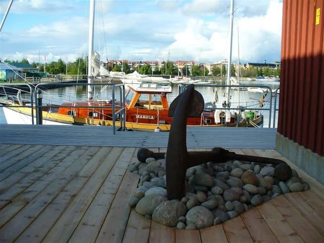 En solbelyst brygga av trä med träplankor och träfläckar sträcker sig ut i vatten. Bakom bryggan syns en campingplats vid Västra Kajen, omgiven av grönska och natur.