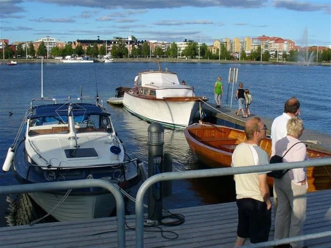 En småbåtshamn vid Västra Kajen camping med flera båtar som ligger förtöjda vid bryggan. Vattnet är stilla och omgivningen grönskande, vilket skapar en fridfull atmosfär.