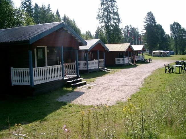 En mysig campingmiljö vid Värnäs Camping med en stuga i trä omgiven av gröna gräsmattor och lummiga träd. Det finns utomhusmöbler och stugan har inslag av ekologiskt boende.