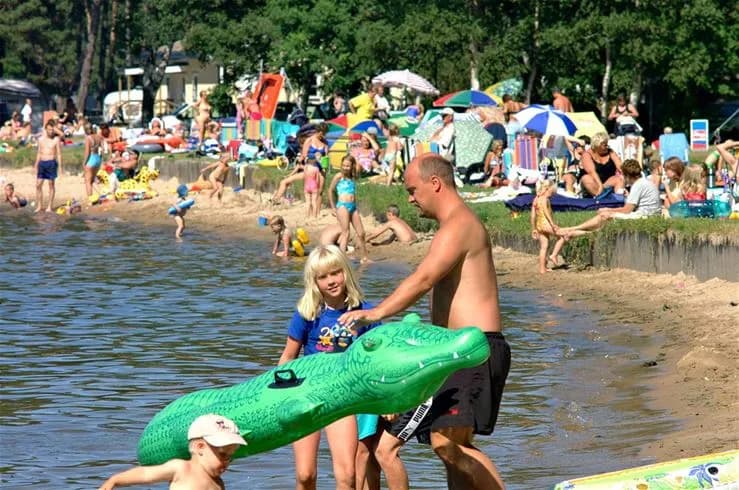 En somrig vy vid Vadstena camping, där människor njuter av sol och bad på en sandstrand. Några klädda i badkläder kopplar av, leker och umgås vid vattenbrynet under en solig semesterdag.