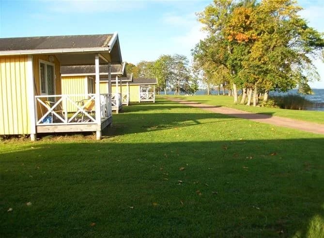 En grönskande campingplats vid Vätterns strand med skuggiga träd och en gräsmatta, perfekt för avkoppling och friluftsliv.