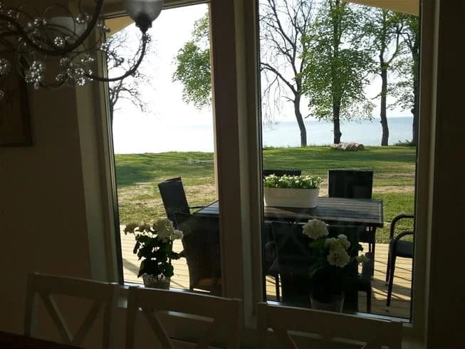 En solig dag vid Vadstena camping med utsikt över kusten. Bilden visar utemöbler inkluderande stolar och ett bord, placerade under en skuggande parasoll nära strandkanten.