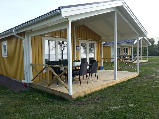 En trästuga med veranda och tak omgivet av en grönskande trädgård på Vadstena camping.