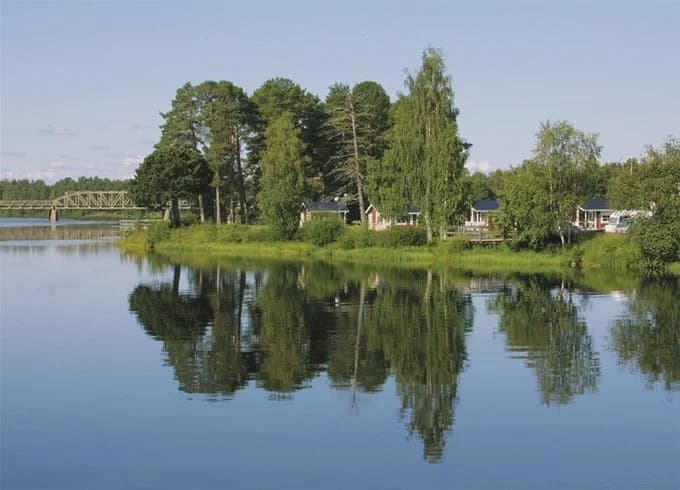 En grönskande campingplats vid vattnet med träd som omger en stilla sjö eller flod, vilket skapar en fridfull och naturnära miljö.