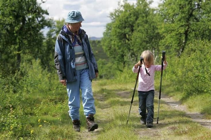 En familj klädd i fritidskläder och med väskor strosar genom en grässlätt vid Svegs camping. I bakgrunden syns grönska och barn som leker i naturen.