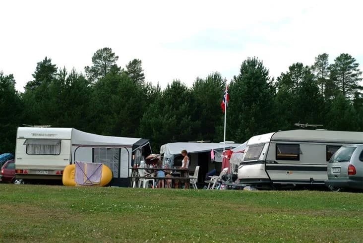 En bild av Strömsunds camping som visar ett grönområde med husbilar, husvagnar och tält uppställda. Flera människor ses njuta av den natursköna miljön på campingen, med gräsytor och parkerade fordon i bakgrunden.