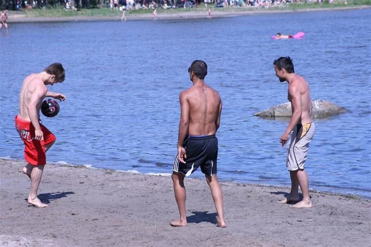 En grupp människor i badkläder njuter av en solig dag på Strandstuvikens havsbad & camping. De går barfota på stranden, omgivna av sand och hav. En känsla av sommar och fritid genomsyrar scenen.