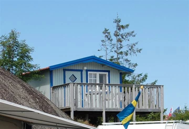 Bild av en pittoresk stuga vid Sotenäs camping, med en veranda prydd med utemöbler. Stugan har en träfasad och en entrédörr som öppnar upp mot en ljus och inbjudande altan omgiven av grönska.