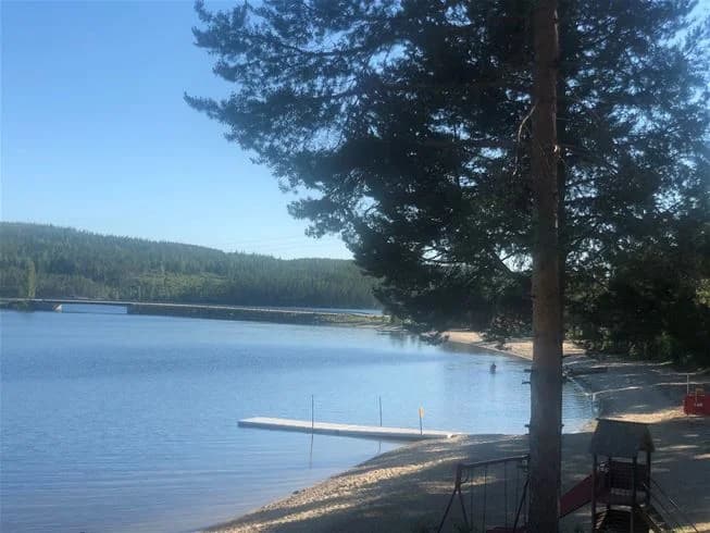 En solig dag vid Sandnäsets bad & camping med en klarblå himmel speglande sig i det stilla vattnet. Förgrunden visar en sjö eller kanal omgiven av grönskande strandlinjer.