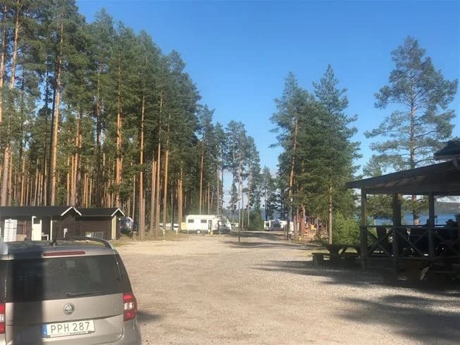 En parkeringsplats vid Sandnäsets bad & camping med flera olika typer av bilar parkerade, inklusive familjebilar och SUV:ar. Området är skuggat och liknar en typisk camping parkeringsplats.