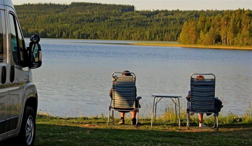 En husbil står parkerad på Saiva Camping & Stugby, omgivet av campingstolar och bord. Solen skiner över området, vilket skapar en inbjudande atmosfär för utomhusaktiviteter och avkoppling.
