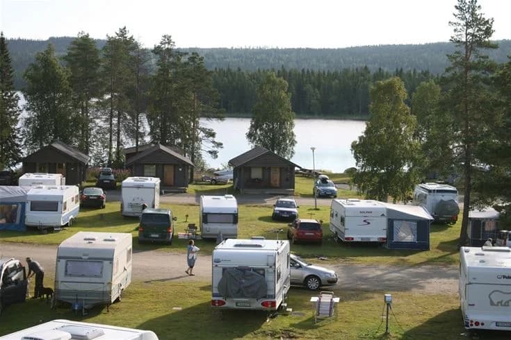 En mysig stuga vid en campingplats, omgiven av en grönskande trädgård och höga barrträd. I bakgrunden syns ett naturskönt område som påminner om Lake District.