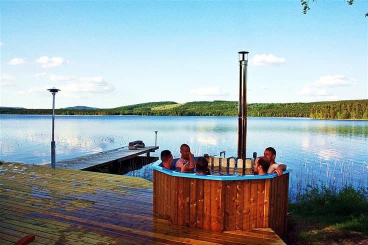En träveranda med utemöbler och en badtunna, omgiven av naturskön miljö vid en sjö på Saiva Camping & Stugby i Lake District.