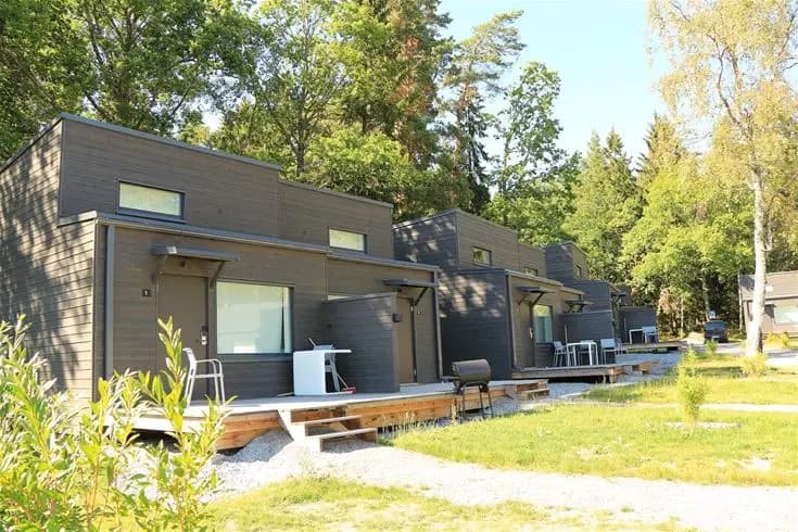 En solig dag vid Rösjöbaden Camping & Stugby, med en pittoresk stuga omgiven av grönskande trädgård och en stenlagd gångväg.