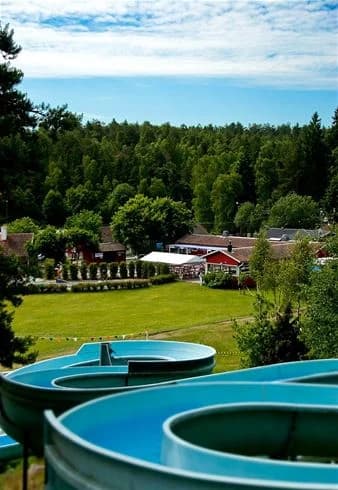En solig dag vid Rösjöbaden Camping & Stugby med besökare som njuter av semesteraktiviteter i en vattenpark och swimmingpool, omgivna av grönska och avkopplande miljö.
