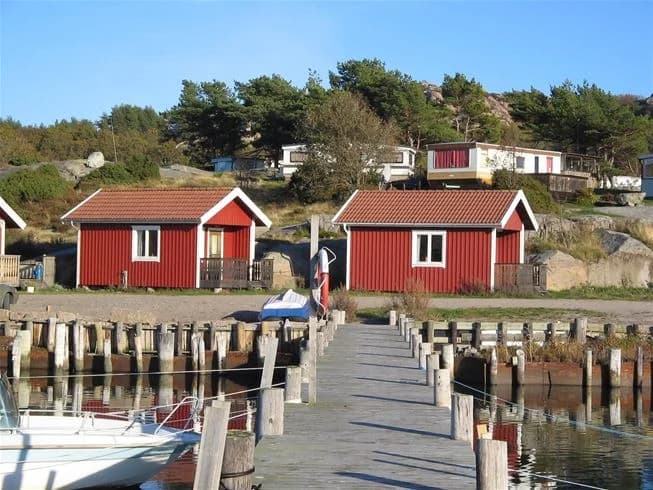 En pittoresk kustbild från Rörviks camping med stugor vid vattnet, småbåtar förtöjda vid en brygga, och en stillsam by i bakgrunden.