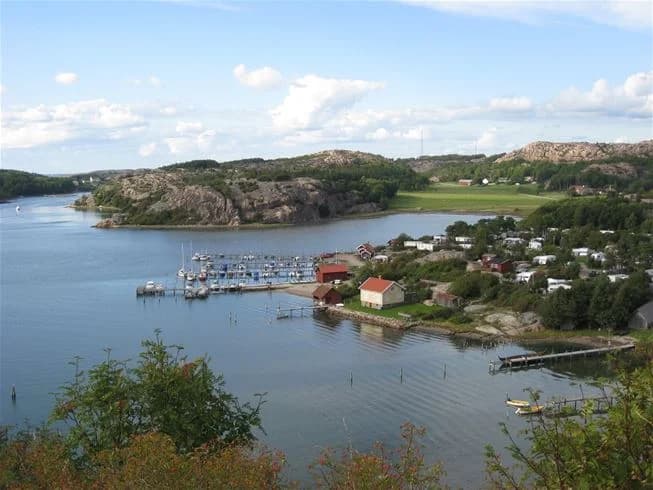 En solig dag vid Rörviks Camping med utsikt över en sjö kantad av gröna banks. Flera båtar ligger förtöjda längs vattnet, omgivna av natursköna landskap och klarblått vatten.