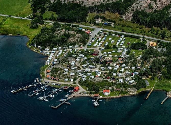 Flygbild över Rörviks camping med kustlandskap: kustlinje, marina, brygga och en närliggande by omgiven av skog.