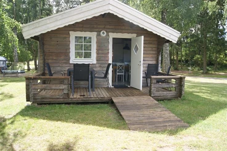 En mysig trästuga med en veranda, omgiven av skuggiga träd vid Orebackens camping. Stugan är byggd i timmer med en mörkbetsad dörr och plankor, perfekt för en avkopplande campingupplevelse.