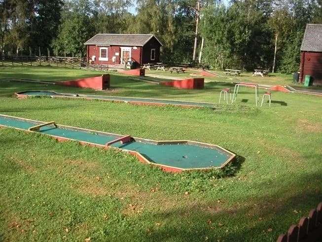 En grönskande gräsyta med en minigolfbana i förgrunden, omgiven av träd och buskar. I bakgrunden syns några personer som ägnar sig åt aktiviteter i ett rekreationsområde på Näsåkers camping & stugby.