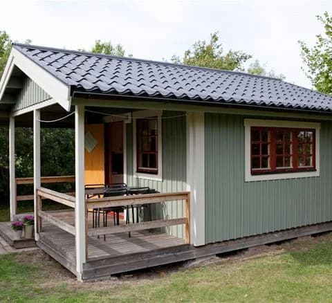 En trästuga vid en campingplats med omliggande trädgård och grönska i skugga.