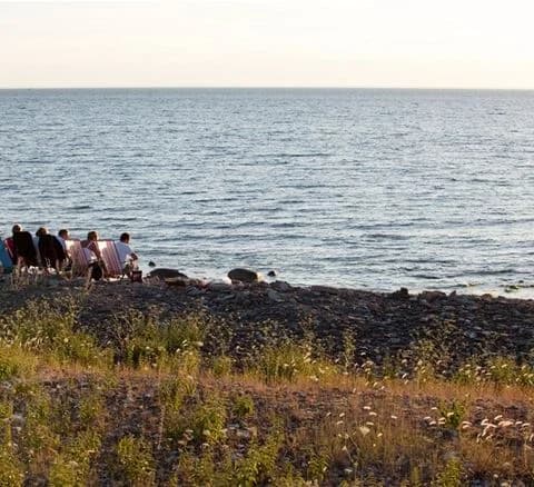 Vy över Neptuni Camping med havet och kusten i bakgrunden. I förgrunden syns klippor och en strand, perfekt för sommaraktivitet och rekreation.