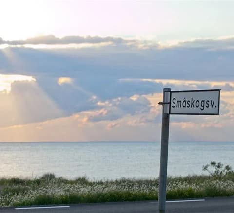 En solig dag vid kusten med en blå himmel och moln vid horisonten. En skylt med gatunamn är synlig i förgrunden, och i bakgrunden syns det glittrande havet.