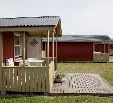 En mysig campingmiljö med en enkel stuga omgiven av grönskande gräsmatta och trädgårdsbyggnader under en klar himmel.