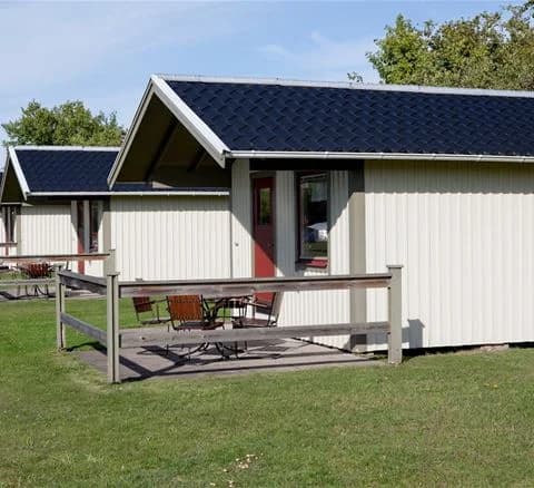 En liten stuga med ett rött tak och vit fasad omgiven av en grön gräsmatta på Neptuni Camping. I bakgrunden syns fler fritidsbyggnader under skuggan av träd.