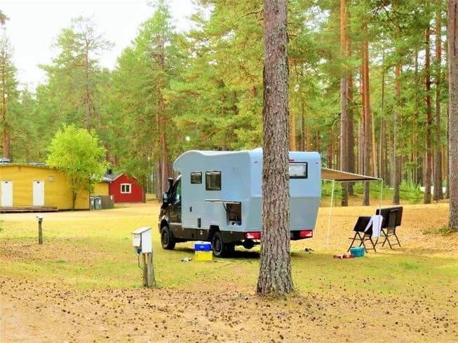 Bild av en naturcamping med husvagnar och husbilar uppställda bland träden. Campingområdet har gröna gräsmattor och skuggiga platser under träden, vilket skapar en fridfull atmosfär i skogsmiljö.