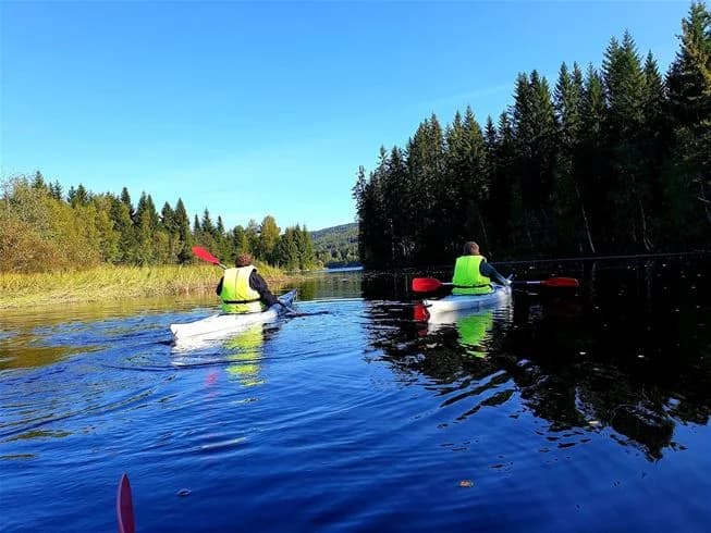 En kajak ligger vid strandkanten av en lugn vattenväg. I bakgrunden syns grönskande skog och blå himmel, vilket skapar en fridfull och inbjudande atmosfär för friluftsaktiviteter.