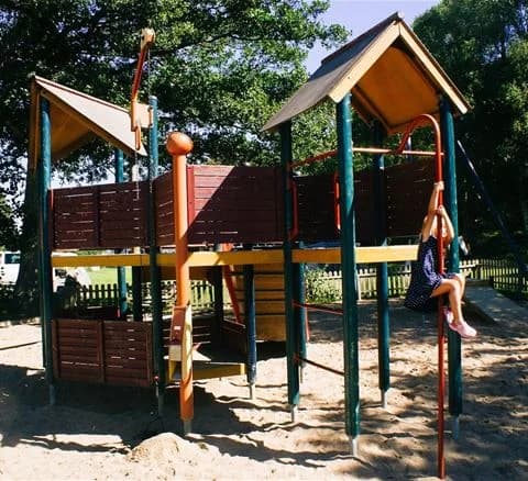 En lekplats på Marstrands familjecamping med olika lekutrustningar, inklusive en rutschkana, omgiven av grönska och solande besökare i ett skuggigt område.