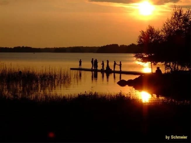En vacker solnedgång vid Mjörknabbens camping, med orange och rosa nyanser som speglas i det stilla vattnet. Naturen omger horisonten, vilket skapar en lugn och idyllisk kvällsstämning.