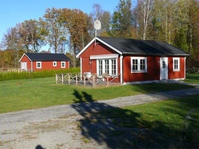 En röd stuga med vita knutar står omgiven av en grön gräsmatta och trädgårdsbyggnader på Mjölknabbens camping.