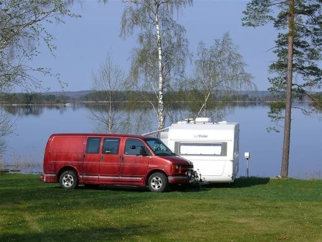 En kompakt husbil står parkerad vid en camping, omgiven av grönska på Mjölknabbens camping. Bilens sidobackspeglar och dörrar syns tydligt.