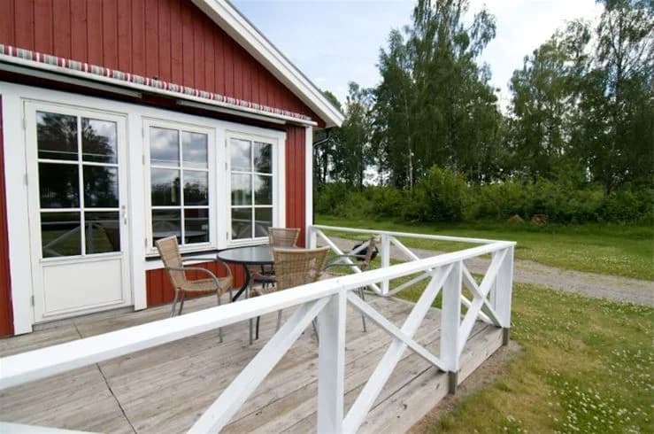 Alt-text: Vy över Mjölknabbens camping med en stuga omgivet av en veranda och trädgård, inhägnad av staket och försedd med utemöbler.