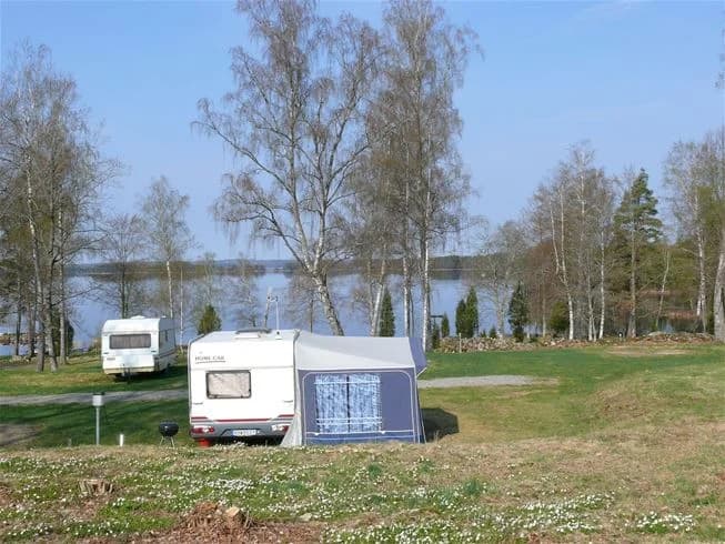 Bild av Mjölknabbens camping med grönt gräs och uppställda campingfordon, inklusive husvagnar och tält. Uteplats med trädgårdsmöbler under en presenning.
