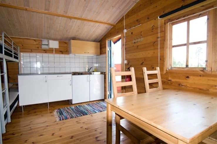 En trästuga med träpaneler och ett trägolv, omgiven av skogsmiljö vid Mjölknabbens camping.