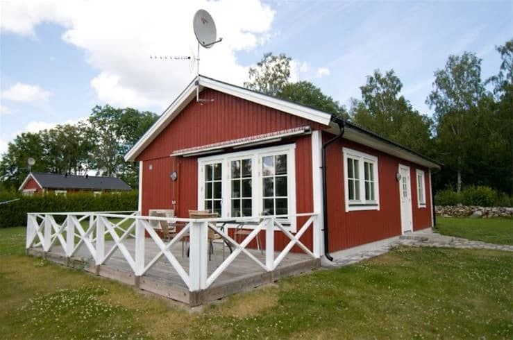 En mysig stuga på Mjölknabbens camping, omgiven av en grönskande trädgård med en veranda, en gårdsplan och omgivande uthus i bakgrunden.