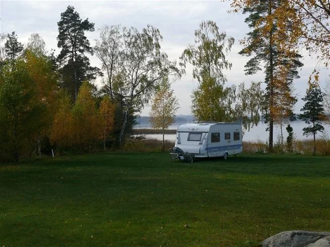 En husvagn står parkerad på en grön gräsmatta vid Mjölknabbens camping, omgiven av träd och nära en sjö.