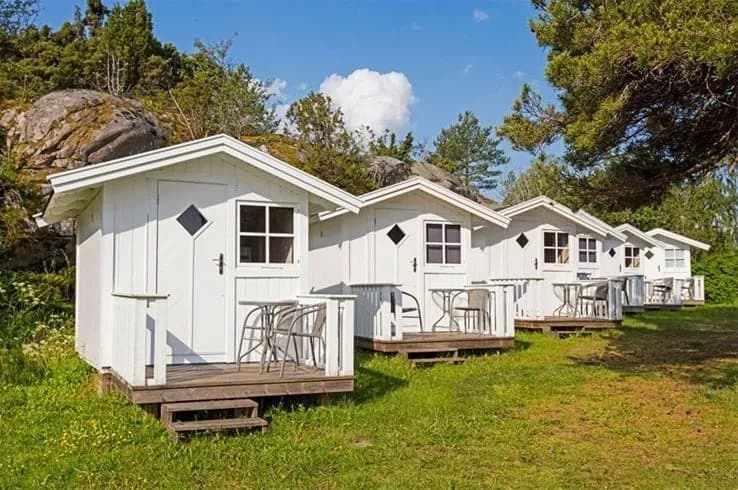 En liten röd stuga med vita knutar omgiven av frodig grönska på Lökholmens camping. I förgrunden syns en välskött gräsmatta.