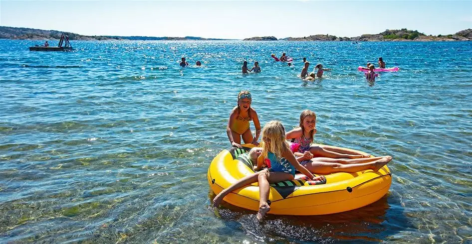 En avkopplande sommardag på Lökholmens camping med utsikt över havet, där besökare njuter av stranden och aktiviteter vid kusten. En båt syns i bakgrunden, vilket förstärker semesterkänslan.