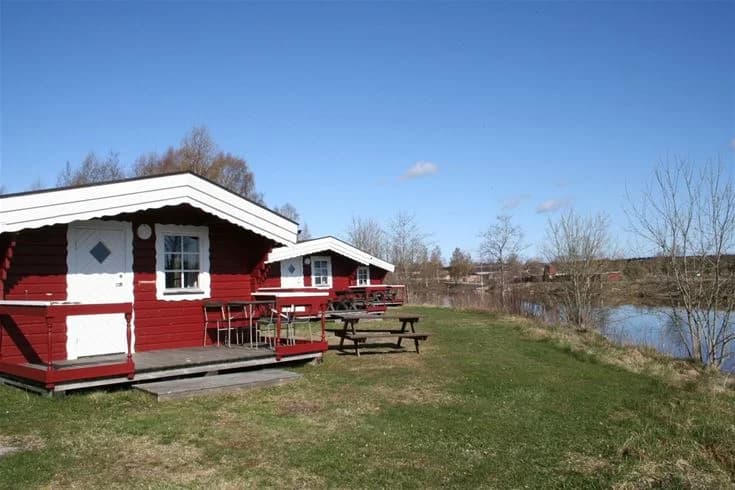 En grön gräsmatta med skuggade områden i en parkmiljö. En timmerstuga syns i bakgrunden på Ljusnefors camping.