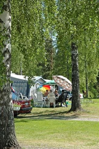 En campingplats med gröna gräsmattor och skuggande träd, där personer njuter av fritiden. Tält är uppställda i bakgrunden, och atmosfären känns lugn och naturnära.