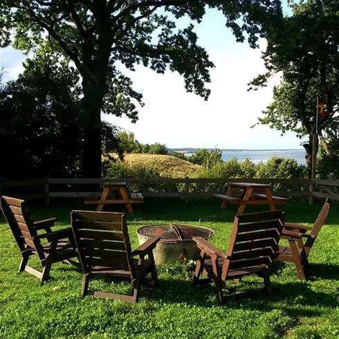 En solig dag på Kiviks familjecamping med trämöbler utomhus, inklusive bord, stolar och bänk i en skuggig trädgårdsmiljö.