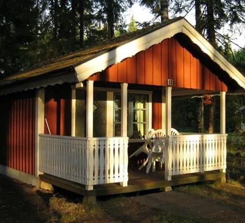 En trästuga med veranda omgiven av grönska, nära Jälluntofta camping. Stugan har ett snedtak och mörkt träpanel som smälter in i den naturliga omgivningen.